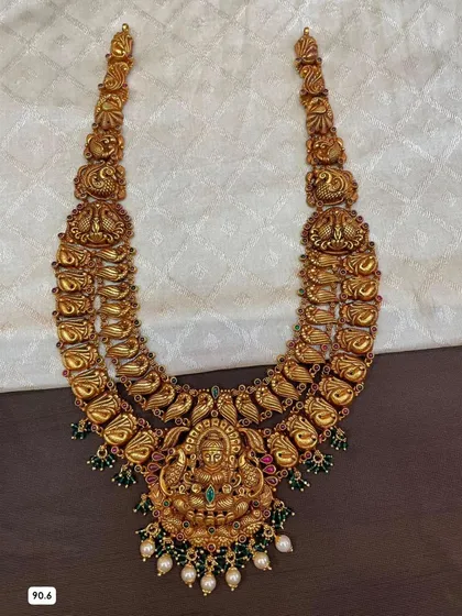 This double-layered temple haram is crafted from 92.5 silver with gold plating. It features intricate peacock and goddess motifs, and is embellished with green stones and pearls.