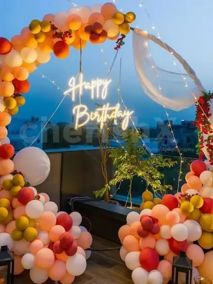 An elegant open-air birthday celebration on a terrace. This setup uses a semi-circular balloon arch in warm sunset tones with a hanging neon sign, sheer drapes, and lanterns, creating a breathtaking view against the evening sky.