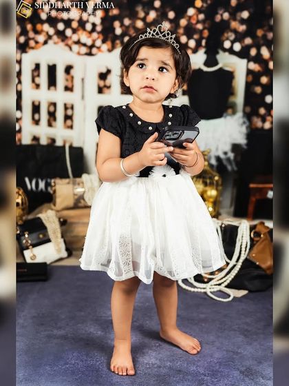 A toddler girl in a black and white dress and a tiara stands confidently in a chic, fashion-themed setup.