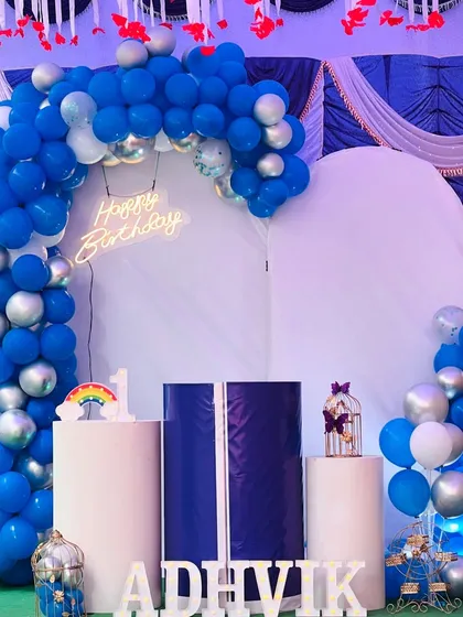 A classic blue and silver theme for a first birthday party. This setup includes an L-shaped balloon garland, a white arched backdrop, and marquee letters for the name 'ADHVIK'.