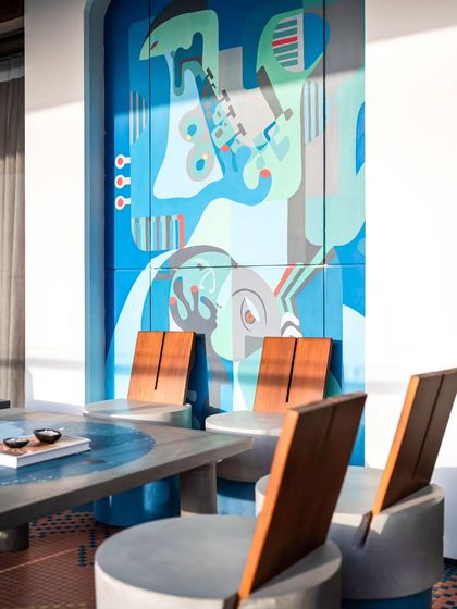 Titled 'Embrace', this large outdoor mural was painted on concrete panels. The abstract figures and cool blue palette create a welcoming and artistic focal point for the seating area.