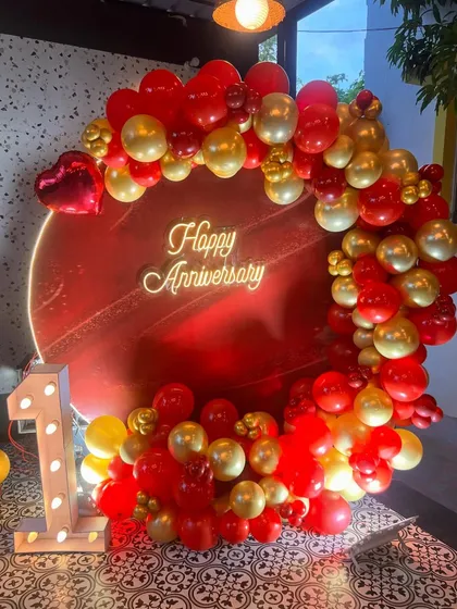 I design bold and beautiful backdrops to celebrate your special milestones. This first-anniversary setup features a vibrant red circular panel, a custom 'Happy Anniversary' neon sign, and a lush garland of red and gold balloons, complete with an illuminated number '1'.
