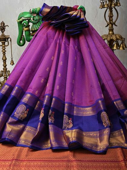 A stunning magenta and royal blue Kanjivaram silk saree. The body features delicate gold stripes and peacock motifs along the border, a classic design that symbolizes grace and beauty.