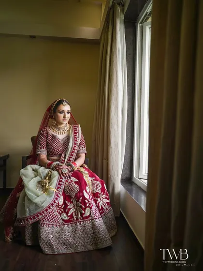 A pensive bridal portrait by the window. The natural light beautifully illuminates the bride in her red lehenga, capturing a moment of calm and anticipation before the ceremony.