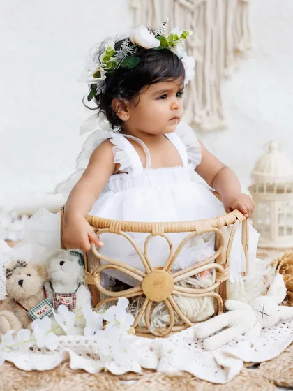 A little angel in a white dress and floral crown sits in a beautiful wicker basket, surrounded by soft, neutral props.