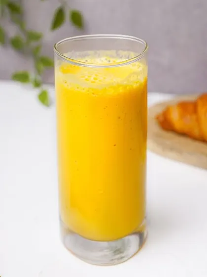 A vibrant orange-colored smoothie in a tall glass, with a croissant and gold spoon creating a pleasant breakfast scene. The bright, natural lighting style is ideal for lifestyle and menu photography.