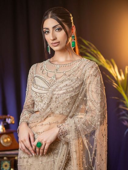 An elegant and sophisticated look suitable for a reception or evening event portfolio. The model wears a delicately embroidered sheer saree, with makeup focused on sharp, defined eyes and a neutral lip, accented by modern emerald jewellery.