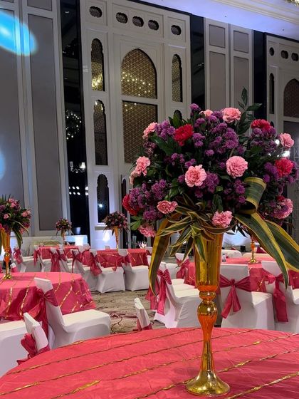 A close up of a tall floral centerpiece from the Mehendi ceremony. The arrangement in a gold vase features a mix of pink and purple carnations and other seasonal blooms.