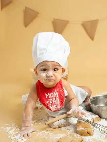 This little baker, with a dusting of flour on his nose and a cute "Mom + Dad = Me" bib, crawls towards the camera, ready for his next kitchen adventure.