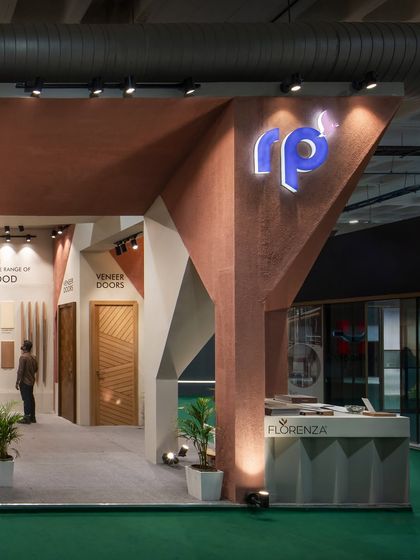 The geometric complexity of the RP Plywood stall is visible from this angle, with faceted surfaces and a strong sense of verticality. The illuminated logo is integrated directly into the architectural form.