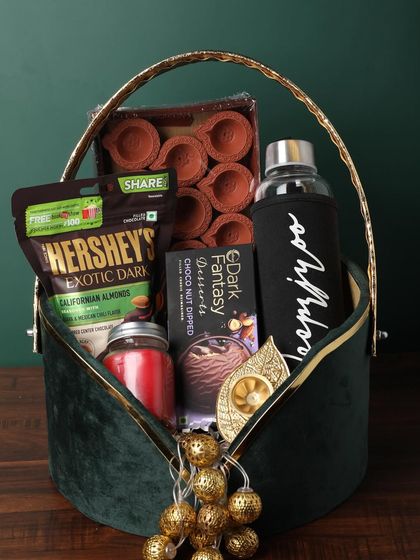A classic Diwali hamper in a green velvet basket. It includes a set of terracotta diyas, a personalized bottle, a scented candle, and a variety of chocolates and nuts.