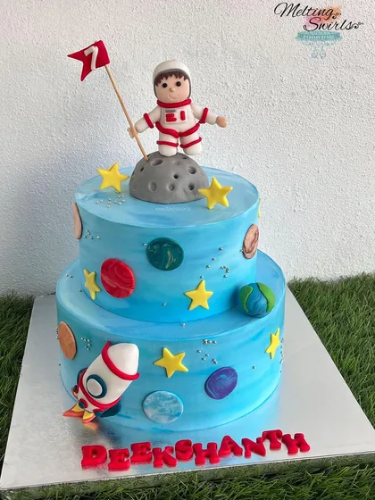 A two-tier space-themed cake for a 7th birthday. This cake is decorated with fondant planets, stars, a rocket, and a handmade astronaut topper.