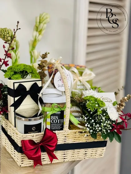 A gourmet gift basket perfect for a birthday or thank you gift. It includes a mix of sweet and savory treats like biscotti, along with a scented candle and fresh flowers, all arranged in a lovely woven basket.