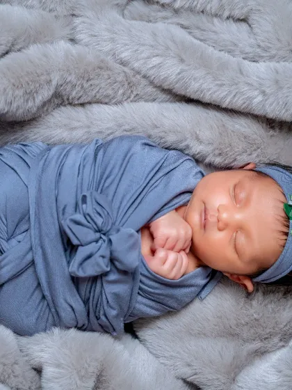 A full-body shot showing the popular "potato sack" wrap pose, which keeps the baby snug and secure. The soft grey fur blanket and matching headband complete this adorable and comfortable setup.