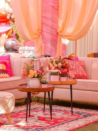 A comfortable and stylish lounge area with plush beige sofas, colorful cushions, and macrame stools, all under a canopy of pink and orange drapes.