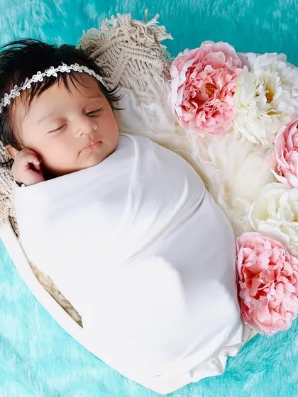 A sleeping baby wrapped in white, nestled in a heart-shaped bowl with pink and white flowers. The aqua-colored blanket adds a pop of fresh color.