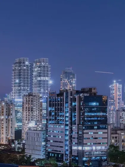 The sleek, modern buildings of the Mumbai skyline are captured at night, with a streak of light from an airplane visible in the sky.