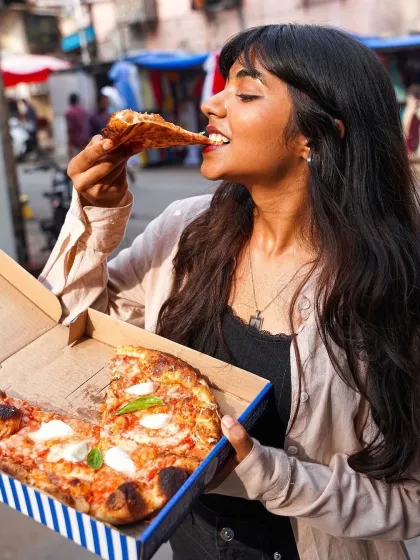 A lifestyle shot of a woman enjoying a slice of pizza in a bustling market setting. This image captures a candid, joyful moment, perfect for a cloud kitchen or takeaway brand wanting to show real-life enjoyment.