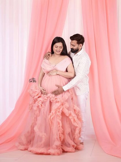 The soft pink backdrop complements the gown perfectly in this studio session. It shows how a simple setup can create a high-end, polished look for a couple's maternity announcement.