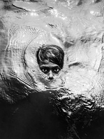 A child's face emerges from the dark water, their eyes wide and captivating in a dramatic and artistic black and white portrait that blurs the line between life and art.