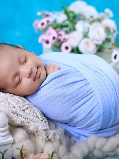 A picture of pure bliss. We are experts at soothing babies and capturing those heart-melting sleepy smiles. This newborn is comfortably swaddled against a soft blue backdrop, surrounded by delicate flowers.