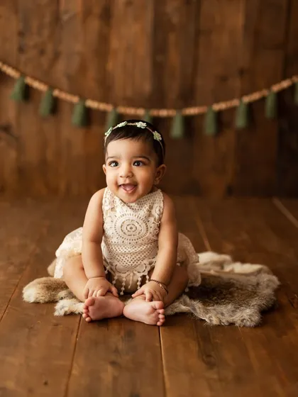 A sweet, cheeky expression from this little one sitting on a fur rug against a rustic wood background.