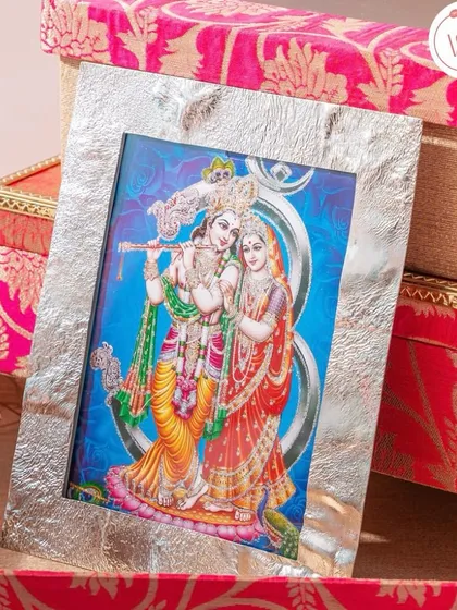 A silver-plated photo frame featuring an image of Radha and Krishna, set against a backdrop of vibrant pink brocade boxes. A divine and thoughtful gift.