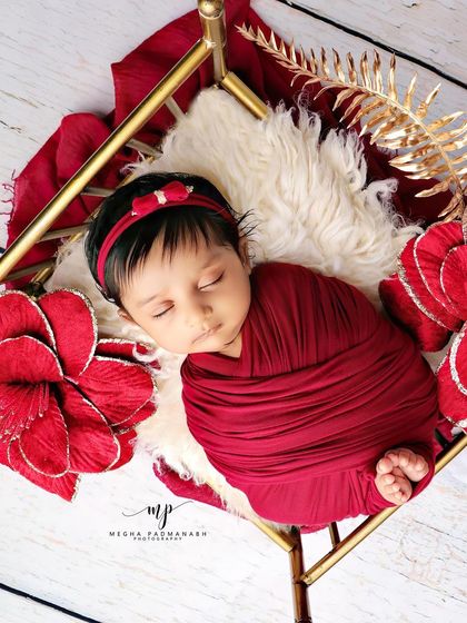 A baby wrapped in rich red, resting on a golden-framed bed with matching red flowers.