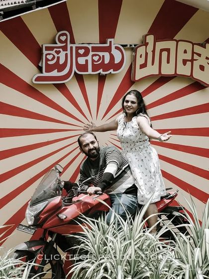 A quirky and fun shot of a couple on a motorcycle in front of a retro-style wall with Kannada script, perfect for a local, vintage-themed shoot.