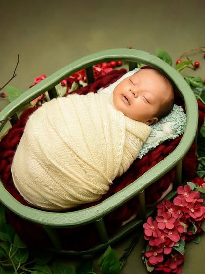 A baby swaddled in cream rests in a basket surrounded by red berries and green foliage. This setup has a lovely, natural, and slightly festive feel.