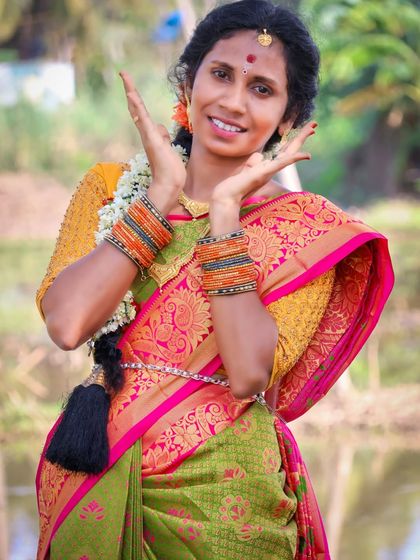 An expressive outdoor portrait of a woman in a traditional green and pink saree. Her playful pose and bright smile are captured against a natural, leafy backdrop.