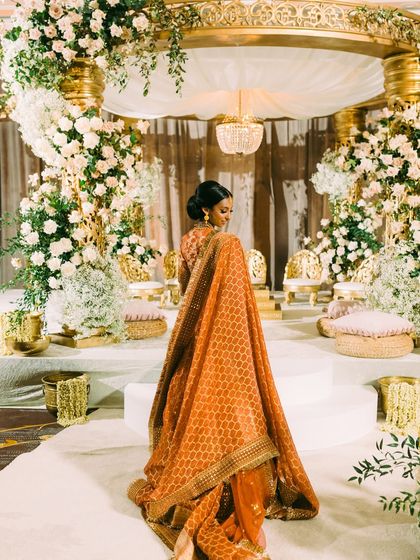 A moment of quiet beauty before the ceremony. The bride, Nisha, stands before the magnificent mandap, her orange Sabyasachi Banarasi silk lehenga a stunning tribute to tradition.