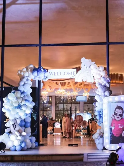 A grand entrance for a teddy bear themed first birthday. The doorway is framed by a blue and white balloon arch, with large, illuminated photo frames on either side displaying pictures of the birthday boy.