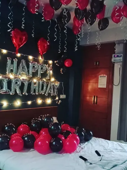 A classic birthday surprise decoration for a bedroom. This setup includes 'HAPPY BIRTHDAY' foil balloons on the wall with fairy lights, and a combination of red and black balloons on the ceiling and bed.