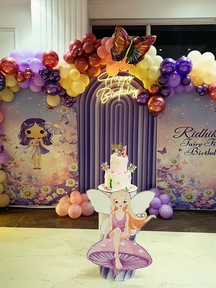 A whimsical 'Fairy First Birthday' setup featuring a custom backdrop with fairy illustrations, butterfly accents, and a pastel balloon garland in purple, pink, and yellow.