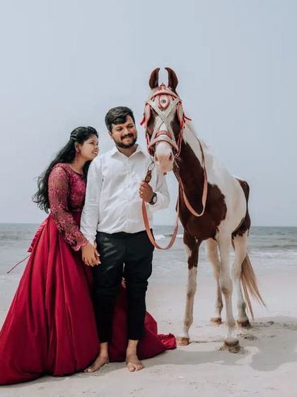 A beautiful pre-wedding portrait on the beach with a horse. The bride's flowing red dress adds a pop of color and drama to the scene.