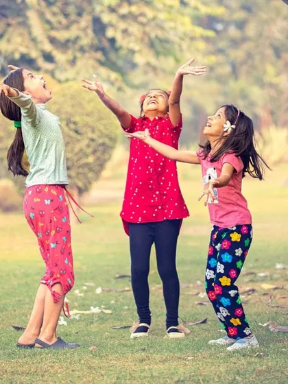 Three young girls play joyfully together in a park, with their arms outstretched towards the sky.