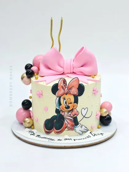 An adorable Minnie Mouse cake featuring a hand-painted illustration, a large pink fondant bow, and balloon decorations.