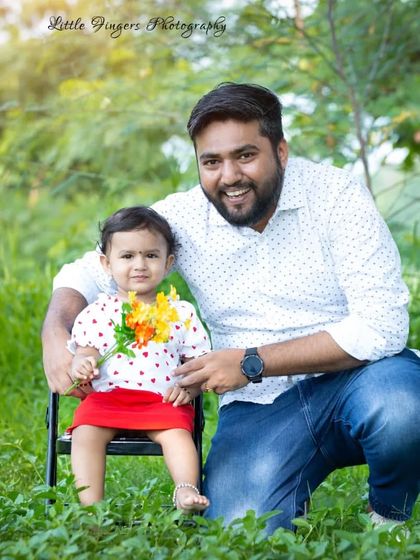 A happy father kneels beside his one-year-old daughter, who is holding a small bouquet of flowers. A bright and cheerful father-daughter portrait.