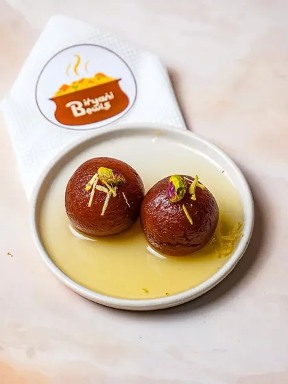 Two gulab jamuns soaked in sugar syrup, garnished with slivered pistachios and served on a white plate with branded tissue. The bright, even lighting makes this classic Indian dessert look irresistible.