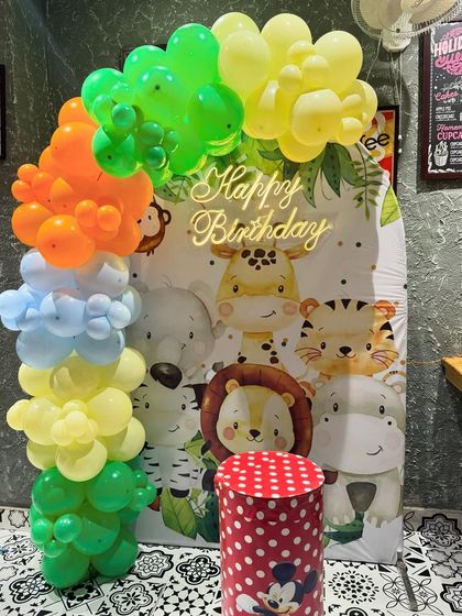 A simple and sweet animal-themed birthday setup. The backdrop features a collection of cute cartoon jungle animals, framed by a colorful balloon garland.