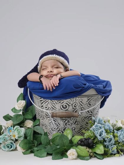 A sleepy baby boy peeks out from a blue swaddle inside a rustic bucket prop. The cascading flowers add a beautiful, gentle touch to this creative newborn setup.