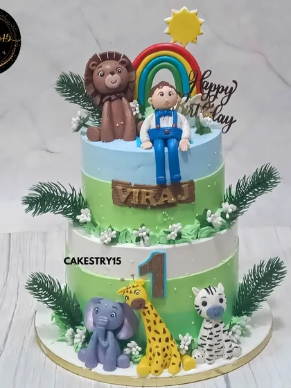 A vibrant two-tier jungle-themed cake for a first birthday. This 3kg choco-vanilla cake is decorated with a boy figurine, a lion, and other cute jungle animals under a rainbow.