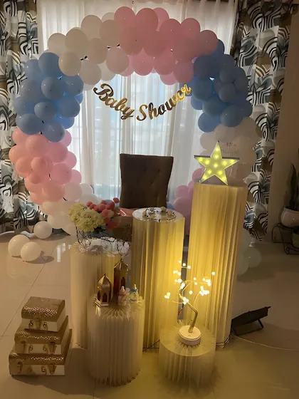 A simple and sweet baby shower setup for an intimate home celebration. The decor includes a balloon arch in pastel pink and blue, illuminated pedestals, and a comfortable chair for the mom-to-be.