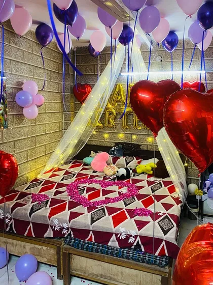 A cozy birthday surprise with purple and pink balloons, large red foil hearts, and a heart made of rose petals on the bed. A fairy light canopy adds a warm glow.