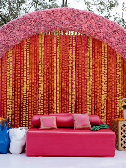 A classic Mehendi seating area featuring a backdrop of orange and yellow marigold strings and a plush pink sofa, perfect for the bride.