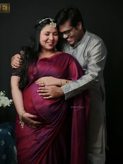 An expecting couple shares a happy moment during their traditional saree maternity shoot. The husband's embrace shows his joy and support.