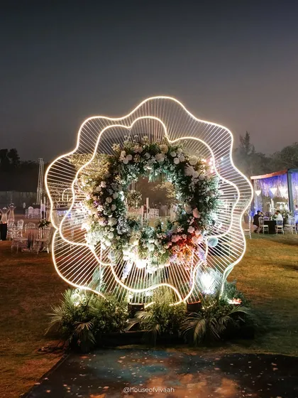 A whimsical and modern photo backdrop made of a neon-outlined, flower-shaped structure filled with a wreath of fresh florals, perfect for a contemporary Sangeet.
