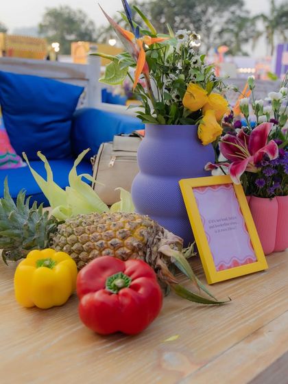 A wide angle of the outdoor carnival setup, showing how fresh fruits and vegetables are used as recurring decor elements on the tables. The vibrant purple and pink vases add pops of color.
