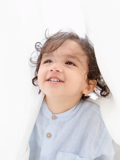 A toddler smiles as he peeks through white curtains, a playful and candid moment from his session.